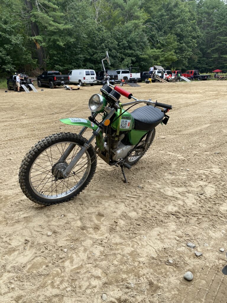 Green Honda dirt bike with a race number plate parked on a sandy area at an outdoor off-road event, with trucks and trees in the background