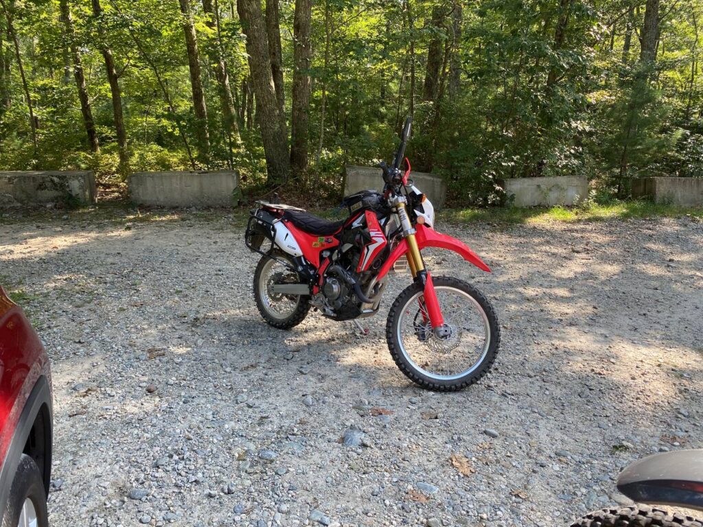 Red Honda CRF 250L with a rear luggage rack, parked on gravel at a wooded trailhead