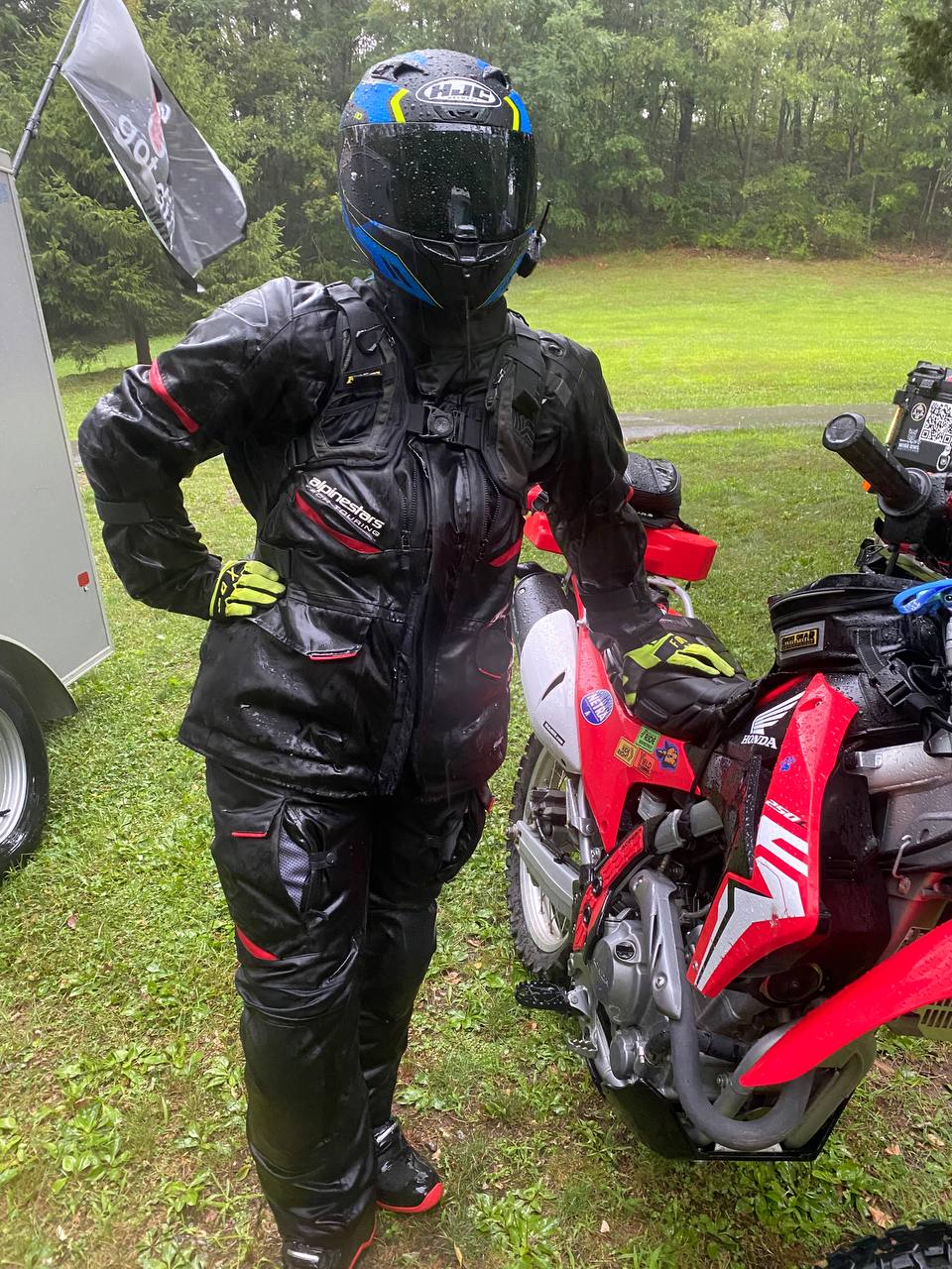 Emily in full rain-soaked riding gear, helmet, armoured jacket and hi-vis gloves, standing beside her red Honda CRF 250L