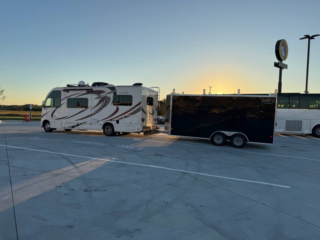 Thor Axis motorhome towing a black Durabuilt enclosed trailer parked at a Buc-ee's at sunrise