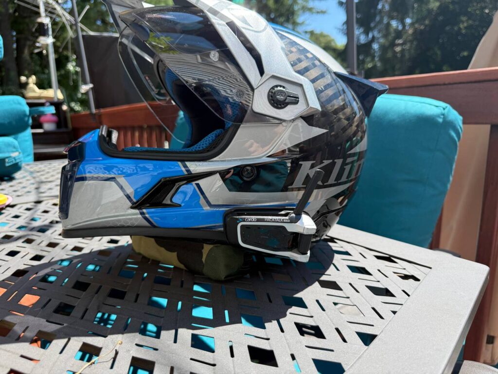 A Cardo Packtalk Bold unit mounted on the side of a blue and white Klim dual sport helmet