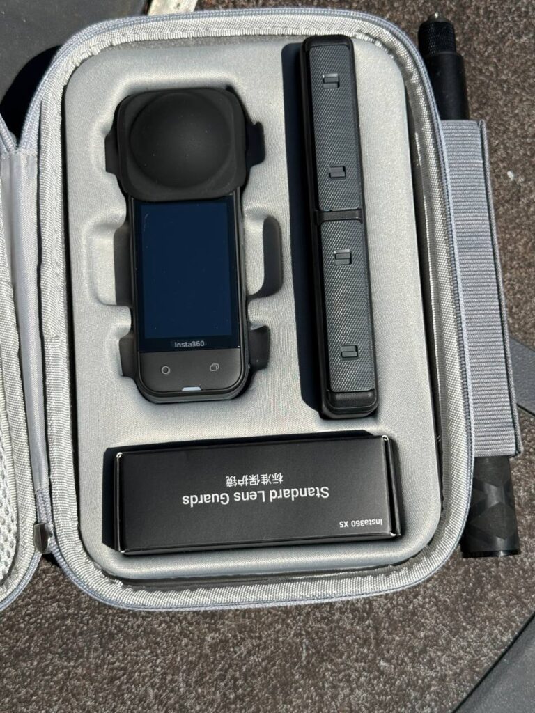 An Insta360 X5 camera in its grey case alongside a selfie stick and a box of standard lens guards
